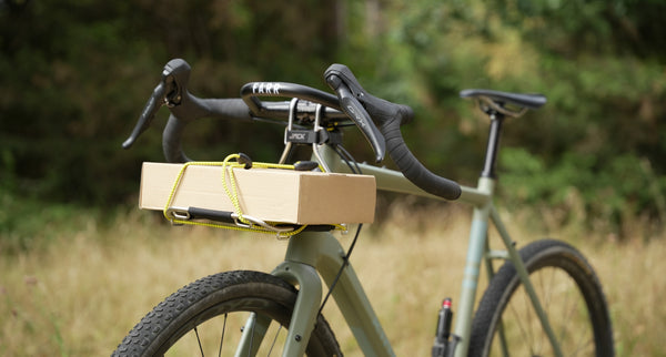 A close-up outdoor shot of a Kinesis AT bicycle with drop handlebars and a cardboard box strapped to the Wholegrain Cycles Jack Classic front rack, with a blurred natural background.