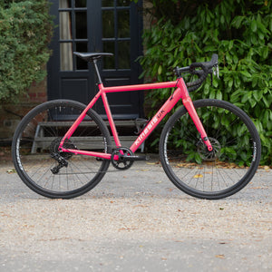 A pink Kinesis UK road bicycle with black wheels and drop handlebars, parked on a gravel surface in front of a black door and green bushes.