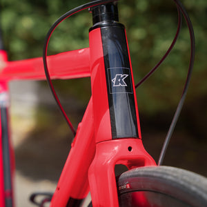 Close-up of a pink bicycle frame with a black vertical section and a white stylised 'K' logo on the head tube, photographed outdoors with blurred green foliage in the background.