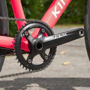 Close-up of a black SRAM Apex bicycle crankset with a chainring and chain, attached to a bright pink road bike frame outdoors.