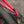 Close-up of a bright pink bicycle frame near the rear wheel, showing a black chain, cassette, brake cable, and a small black logo transfer on the frame. The background is blurred gravel outdoors.