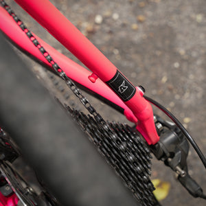Close-up of a bright pink bicycle frame near the rear wheel, showing a black chain, cassette, brake cable, and a small black logo transfer on the frame. The background is blurred gravel outdoors.