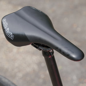 Close-up of a black Selle Italia bicycle saddle mounted on a dark seat post, photographed outdoors against a blurred gray pavement background.