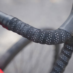Close-up of a bicycle drop handlebar wrapped in black grip tape with small white reflective dots, photographed outdoors against a blurred gray background.
