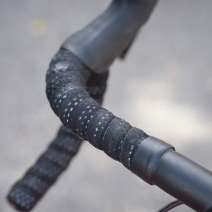 Close-up of a bicycle drop handlebar wrapped in black textured tape with small white dots, set against a blurred gray pavement background.