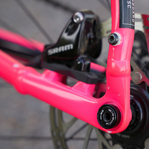 Close-up of a bright pink bicycle frame with a black SRAM brake mech in an outdoor setting.