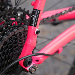 Close-up of a bright pink bicycle frame near the rear cassette, with a black and white Kinesis 'K' logo decal on the down tube and a chainring visible.