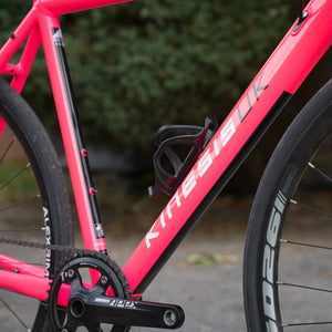 Close-up of a bright pink and black KINESIS UK 4 seasons road bicycle frame, showing the down-tube with white KINESIS branding, a black water bottle, and part of the drivetrain against an outdoor blurred background.