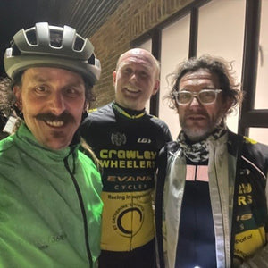 Three men, two of whom are wearing cycling gear, standing together indoors.