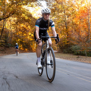 A cyclist wearing a black and blue Scott jersey, black shorts, and a white helmet rides a road bike on an autumn forest road, with another rider blurred in the background. Using Boyd Cycling 36mm Carbon road wheels.