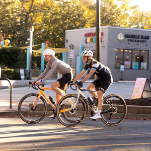 Two cyclists riding road bikes on a city street in front of a storefront, with one person wearing a gray long-sleeve top and black shorts on an orange bike, and the other wearing a black and blue cycling kit on a silver bike. Both are using Boyd Cyling 36mm carbon wheels.