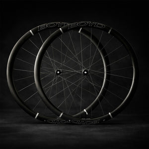 Two black BOYD Cycling 36mm Carbon bicycle wheels with thin black spokes, arranged in a stacked, overlapping composition against a dark, moody studio background.