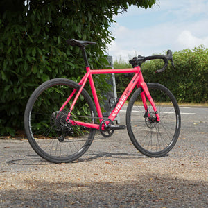 A Kinesis 4S ex-demo pink road bicycle with black components and drop handlebars, parked on a paved path outdoors with green bushes and a blue sky in the background. 