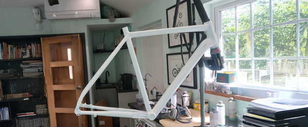 A white bicycle frame displayed indoors on a stand in a room with a window, shelves, and desk items.