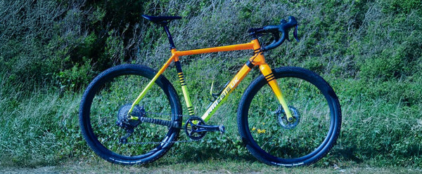 A Kinesis Tripster bicycle with a bright orange and yellow frame, black wheels, and black components, photographed outdoors against a grassy, brushy background.