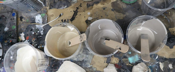 Three plastic buckets filled with light gray and beige liquid paint, each with a wooden stir stick, arranged on a messy, paint-splattered workbench ready to paint Jo Burt's Tripster.