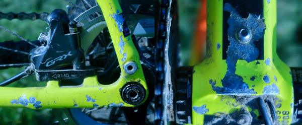 Close-up of a bright neon yellow Kinesis Tripster bike frame with scratches and chips, on the chainstay and bottom bracket area and drivetrain against a blurred green outdoor background.