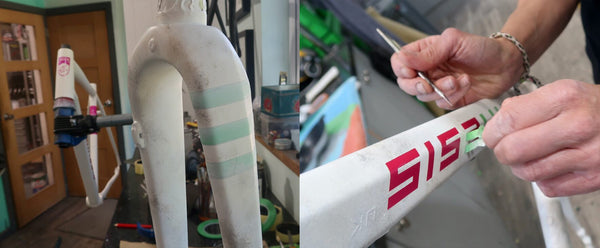 A close-up of Jo's Kinesis Tripster white bicycle fork with green and white stripes, alongside a person's hands applying red lettering to a white component in a workshop setting.