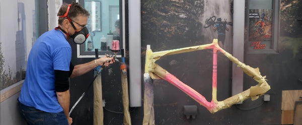 Jo Burt wearing a blue shirt, jeans, and a respirator mask uses a spray gun to paint his Kinesis Tripster bicycle frame. The frame is suspended vertically, showing yellow tape and pink paint on a gray workshop wall.