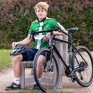 Joshua Moodie with curly blond hair sits on a bench outdoors, wearing a green and white cycling jersey, dark shorts, and holding a black Kinesis Tripster AT Plus bicycle next to him. He is also holding a black cycling helmet.
