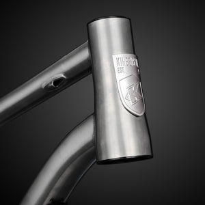 Close-up of a titanium bicycle frame with a branded head badge on a dark background