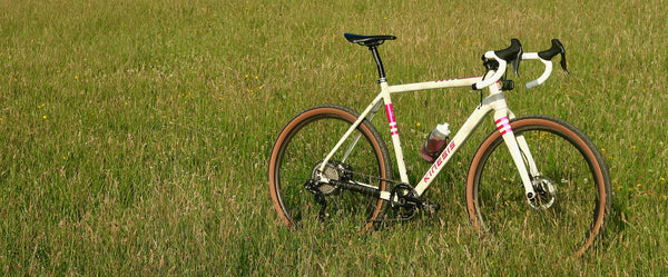 A white gravel bicycle with pink accents and brown sidewall tires is parked on a green grassy field.
