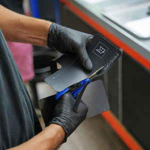 A close-up of a person wearing black gloves using blue-handled scissors to cut a black flexible mudguard flap with a printed logo and text, set against a blurred indoor background.