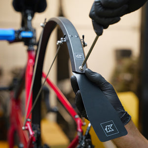 A close-up of a person wearing black gloves using an Allen wrench to put a black mudguard flap on to a Fend Off mudguard against a blurred indoor background.
