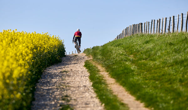 Kinesis ambassador Jo Burt cycling on a gravel path with yellow flowers and a wooden fence on a clear day.