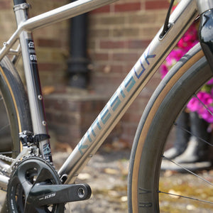 Close-up of a Kinesis Titanium GTD bicycle frame with blue 'KINGSBEEK' branding, a black crankset, and a dark gray wheel, photographed outdoors against a blurred brick wall and pink flowers.