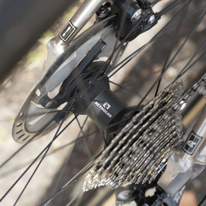 Close-up of a bicycle rear wheel showing a black Reynolds hub, silver cassette, disc brake, and part of the frame with a blurred outdoor background.