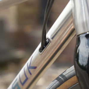Close-up detail of a Kinesis Titanium GTD frame with a black cable running through a cable guard, showing part of the silver metallic down tube and a blurred outdoor background.