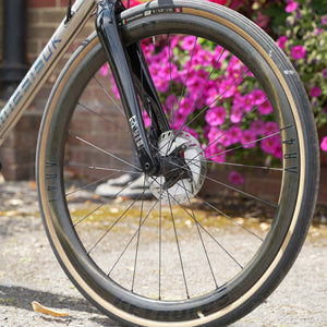 Close-up of a Kinesis Titanium GTD wheel and front fork outdoors, with a black carbon fiber wheel, silver disc brake, and pink flowers blurred in the background.