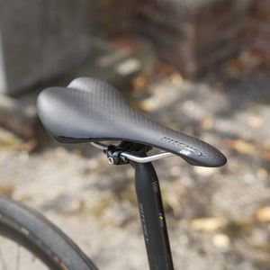 Close-up of a black bicycle saddle mounted on a black bike seat post, photographed outdoors with a blurred gravel and brick background.