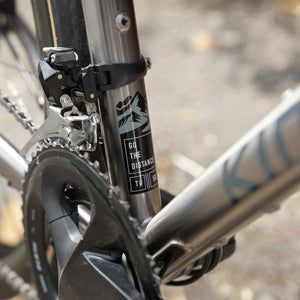 Close-up of a Kinesis Titanium GTD frame with a black seat-tube clamp holding a black saddle, on a mountain bike with a silver fork and black crankset. A black label on the fork reads 'G0 THE DISTANCE'.