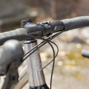 Close-up of a bicycle handlebar with a Ritchey brand black stem and bar-end saddle, mounted on a silver frame with black cables, photographed outdoors with a blurred natural background.