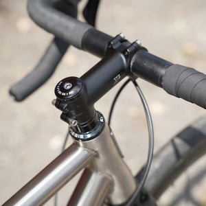 Close-up of a bicycle handlebar and stem assembly, showing black drop handlebars with textured grips, a black stem and headset, and part of a silver metallic frame against an outdoor blurred background.