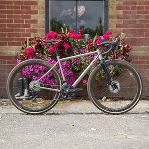 A Kinesis Titanium GTD bicycle with a matte silver-gray frame and black components is parked on a paved street in front of a brick wall and a large planter of bright pink flowers.