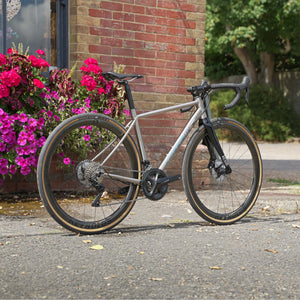 A Kinesis Titanium GTD road bicycle with black components parked on a paved street in front of a brick building with pink flowers.