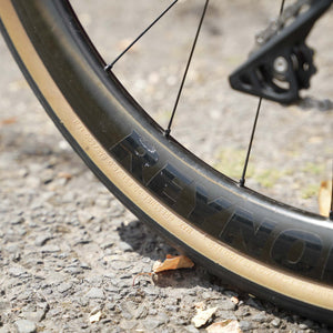 Close-up of a black bicycle wheel with a tan sidewall and embossed REYNOLDS lettering, photographed outdoors on gravel.