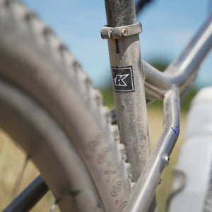 Close-up of a Kinesis Fernweh bicycle frame near the rear wheel, showing a black and white 'K' logo decal on the seat tube. The bike frame is metallic silver, covered in light dust, with a muddy tyre in the foreground and a blurred outdoor background.