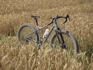 A Kinesis Fernweh  mountain bike with black drop handlebars and knobby tires, parked in a field of dry golden wheat.