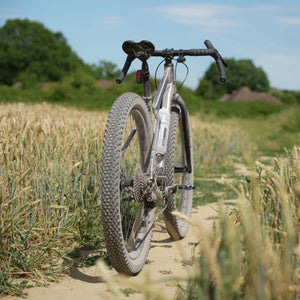 A Kinesis Fernweh MTB bicycle with black handlebars and knobby tires, positioned on a dirt path surrounded by a wheat field under a blue sky.
