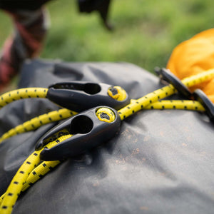 Close-up of yellow and black Wholegrain Cycles bungee with black plastic quick-release buckles resting on a dark grey bag outdoors.