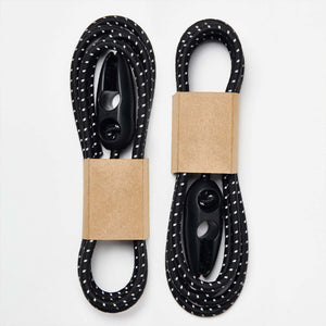 Two Wholegrain Cycles black and white bungees with white speckled braided cords and black plastic jump rings, each wrapped with a brown kraft paper band, on a light grey background.