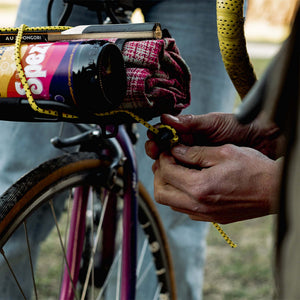 Close-up of a person's hands securing a Wholegrain Cycles Bungee, with a purple bicycle wheel and frame visible in the background.