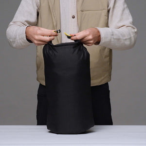 A person wearing a beige vest and light sweater holds open a Wholegrain Cycles black waterproof stuff sack with yellow zipper pulls, standing behind a white table against a grey background.