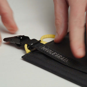 Close-up of a black woven fabric pouch with a black strap and yellow ring, held by two hands. The strap has the brand name 'WHOLEGRAIN' printed on it.