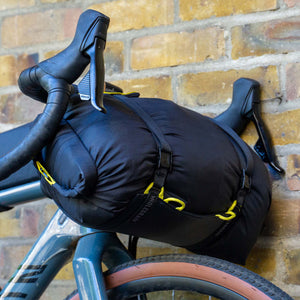 A black Wholegrain Cycles bicycle pannier bag with yellow accents strapped to a bicycle's frame, leaning against a yellow brick wall.
