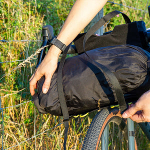 A close-up outdoor shot of a person securing a Wholegrain Cycles black bicycle pannier bag with straps to a bike rack on a bicycle, with grass and a wire fence in the background.